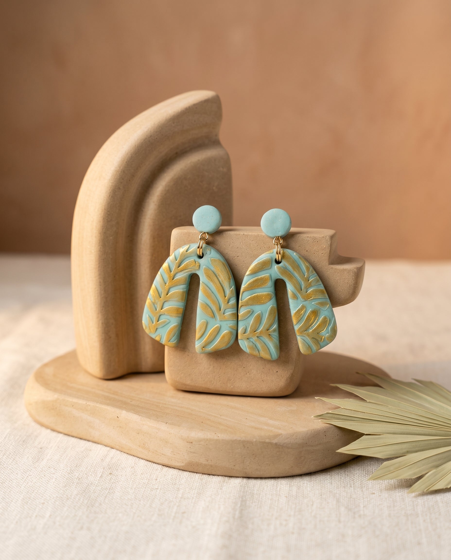 Pendientes Kaiora Bloom de arco azul menta con patrón de hojas doradas, estilo artístico.