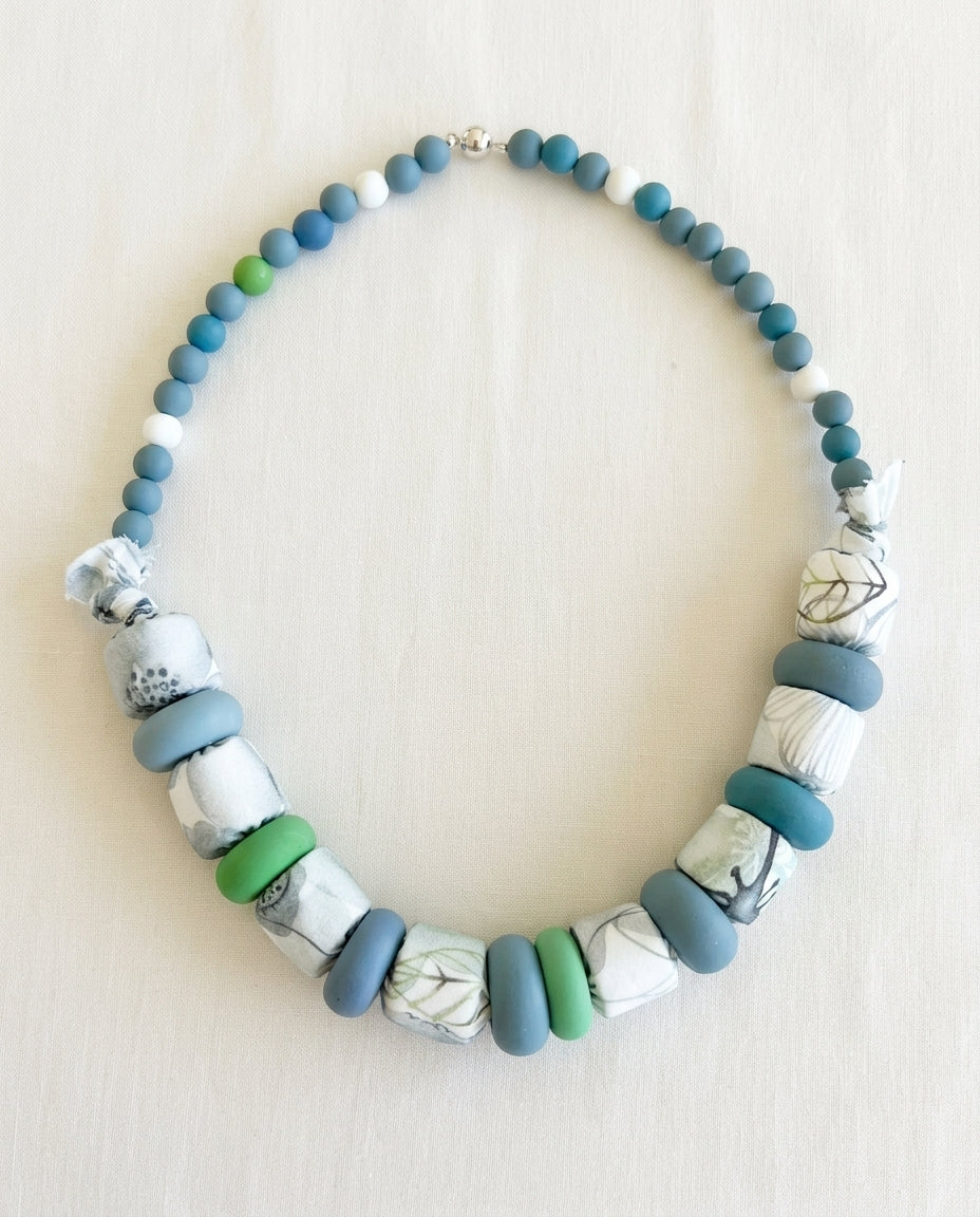 Collar Kaiora Lago artesanal con cuentas textiles botánicas y lisas en tonos azul, verde y blanco.